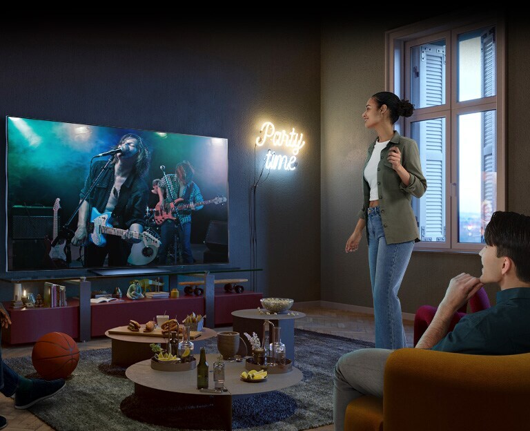 Auf dem TV-Bildschirm läuft ein Konzert, zwei Männer sitzen auf dem Sofa und auf einem Sitzsack, während eine Frau stehend die Musik genießt. Auf dem stehen diverse Snacks wie Popcorn, Nachos und Cola, es handelt sich also um einen richtig schönen Konzertabend.